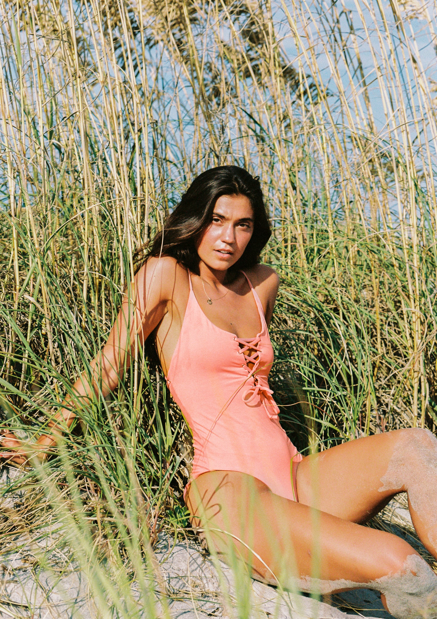 Girl wearing a salmon colored one piece swimsuit at the beach sitting on sand in front of marram grass